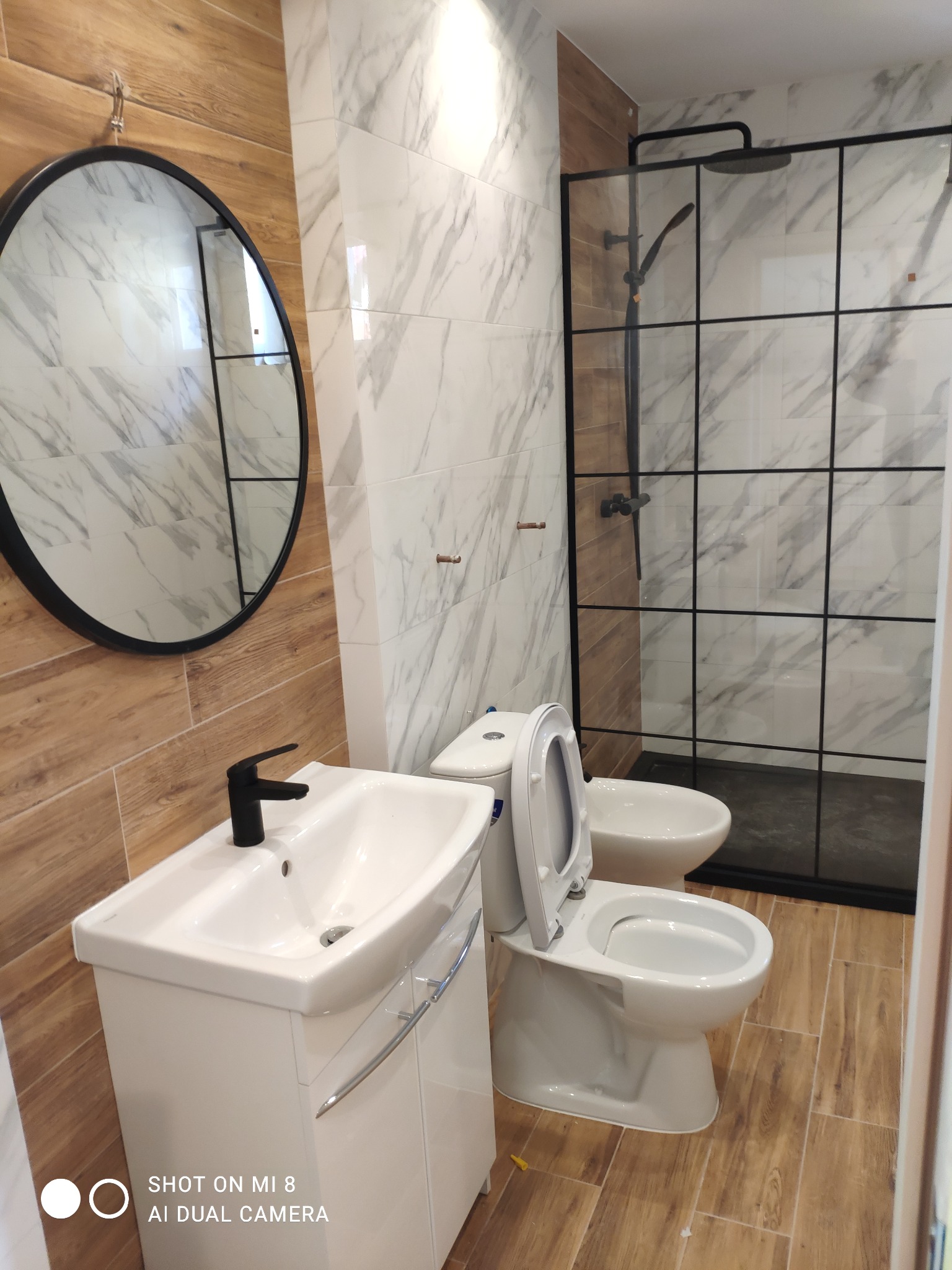 Nowoczesna łazienka z drewnopodobnymi i marmurowymi płytkami, czarną armaturą, okrągłym lustrem i kabiną prysznicową typu walk-in z czarnymi szprosami.