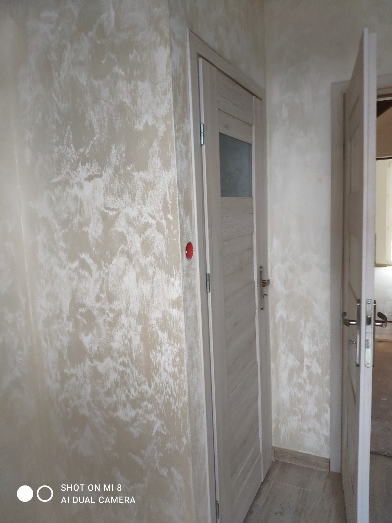 Ściana z efektem dekoracyjnym przypominającym chmurki, widoczne dwie drewniane, pomalowane na jasno drzwi z metalowymi klamkami, jedna zamknięta z małym przeszkleniem, druga uchylona.