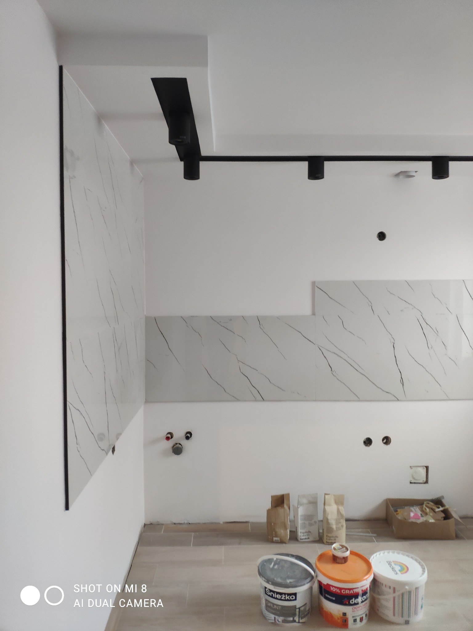 Montaż płytek imitujących marmur na ścianie w kuchni, widoczne puszki z farbą Śnieżka i Dekoral oraz worki z zaprawą, czarne reflektory sufitowe.
