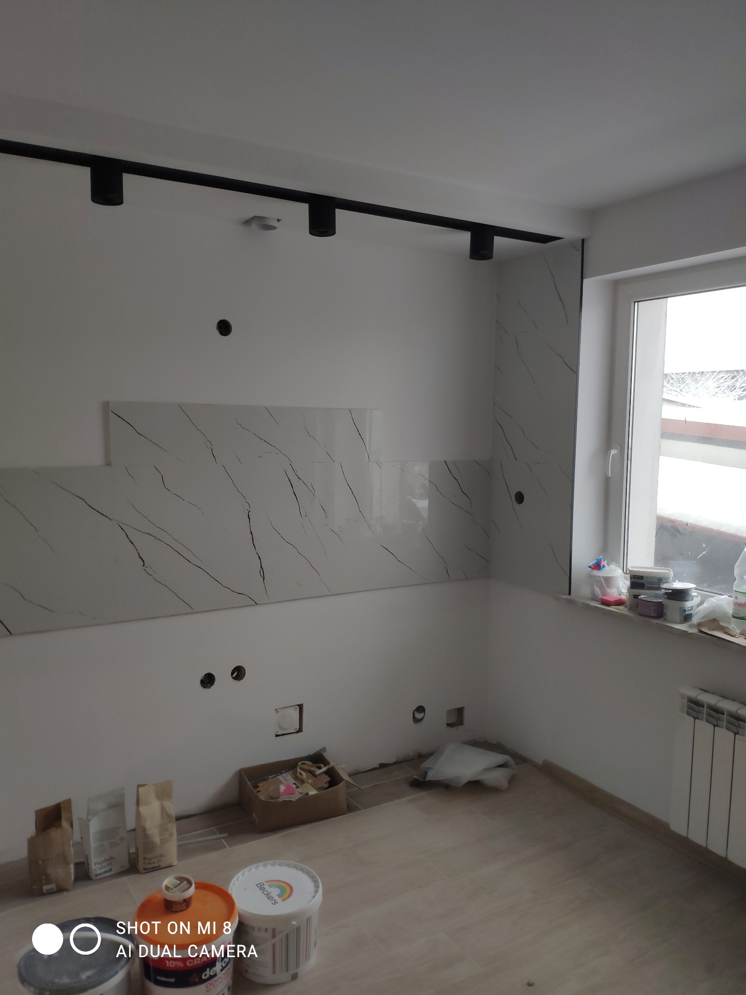 Wnętrze kuchni w trakcie prac wykończeniowych, widoczne płytki imitujące marmur na ścianie, sufit z oświetleniem szynowym, przygotowane materiały budowlane na podłodze, okno z parapetem.