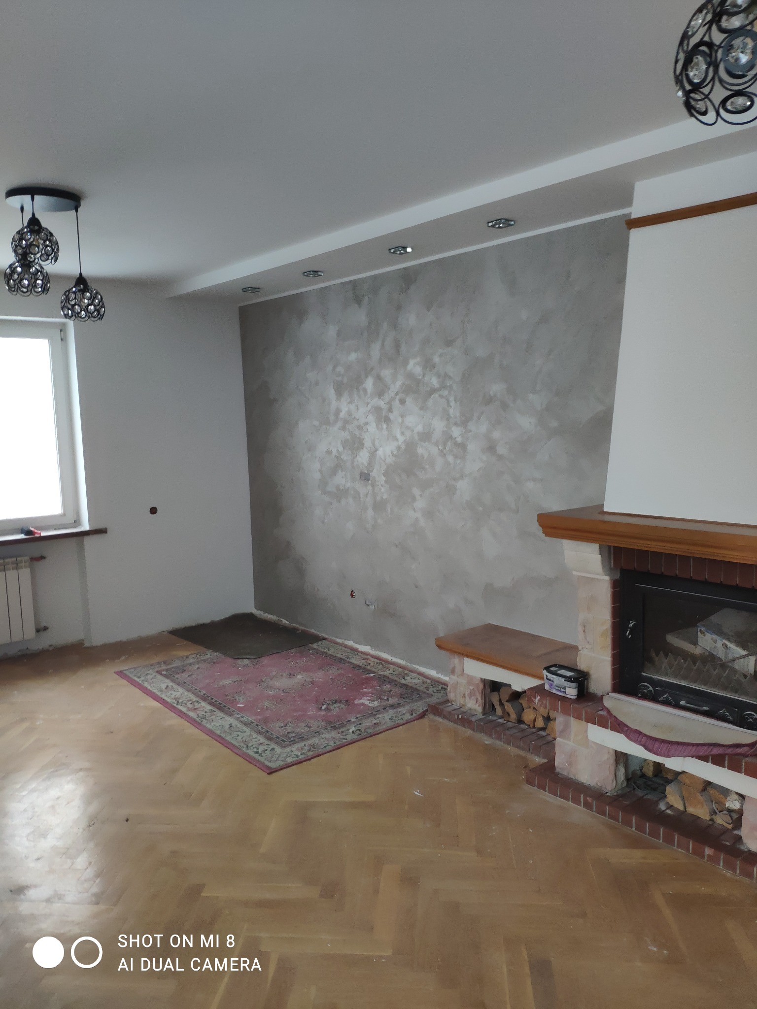 Pokój po remoncie z dekoracyjną gładzią na ścianie za kominkiem, drewnianą podłogą parkietową i dywanem w odcieniach czerwieni