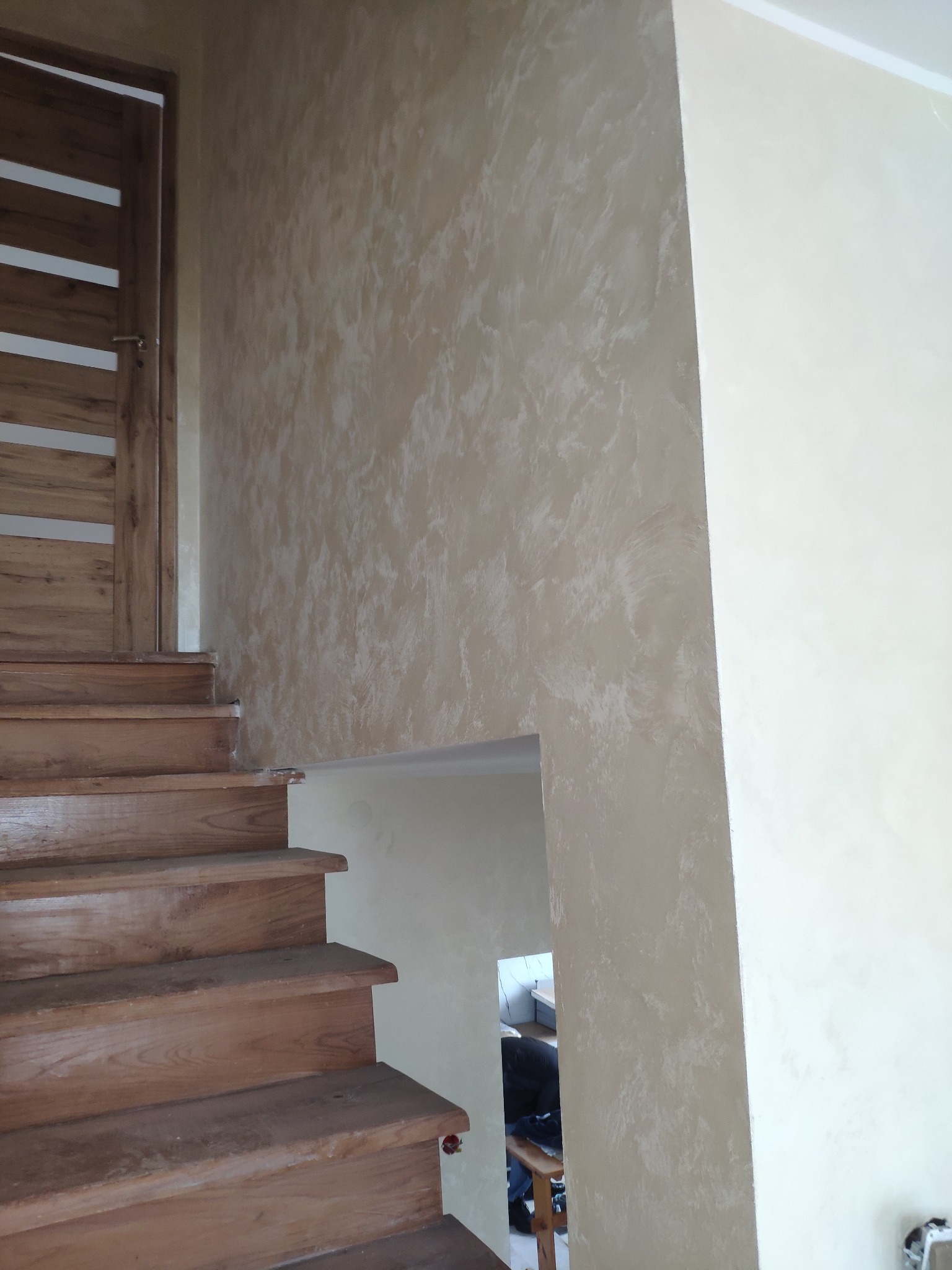 Ściana wykończona dekoracyjną gładzią w odcieniach beżu i bieli, widoczna obok drewnianych schodów i drzwi z przeszkleniami, z fragmentem pomieszczenia w tle.
