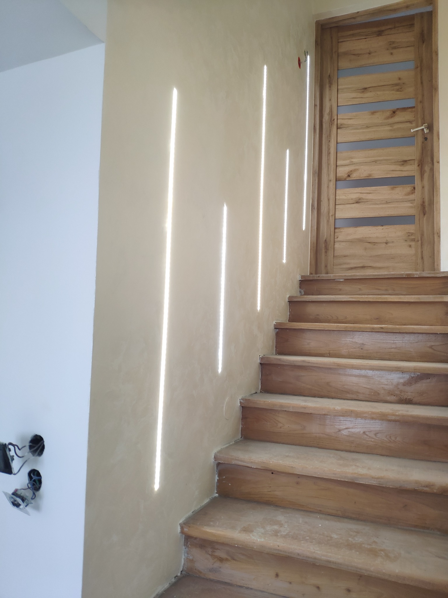 Schody drewniane z efektem światła na ścianie w wykończonym wnętrzu, widoczna gładź na ścianie i drewniane drzwi z przeszkleniami.