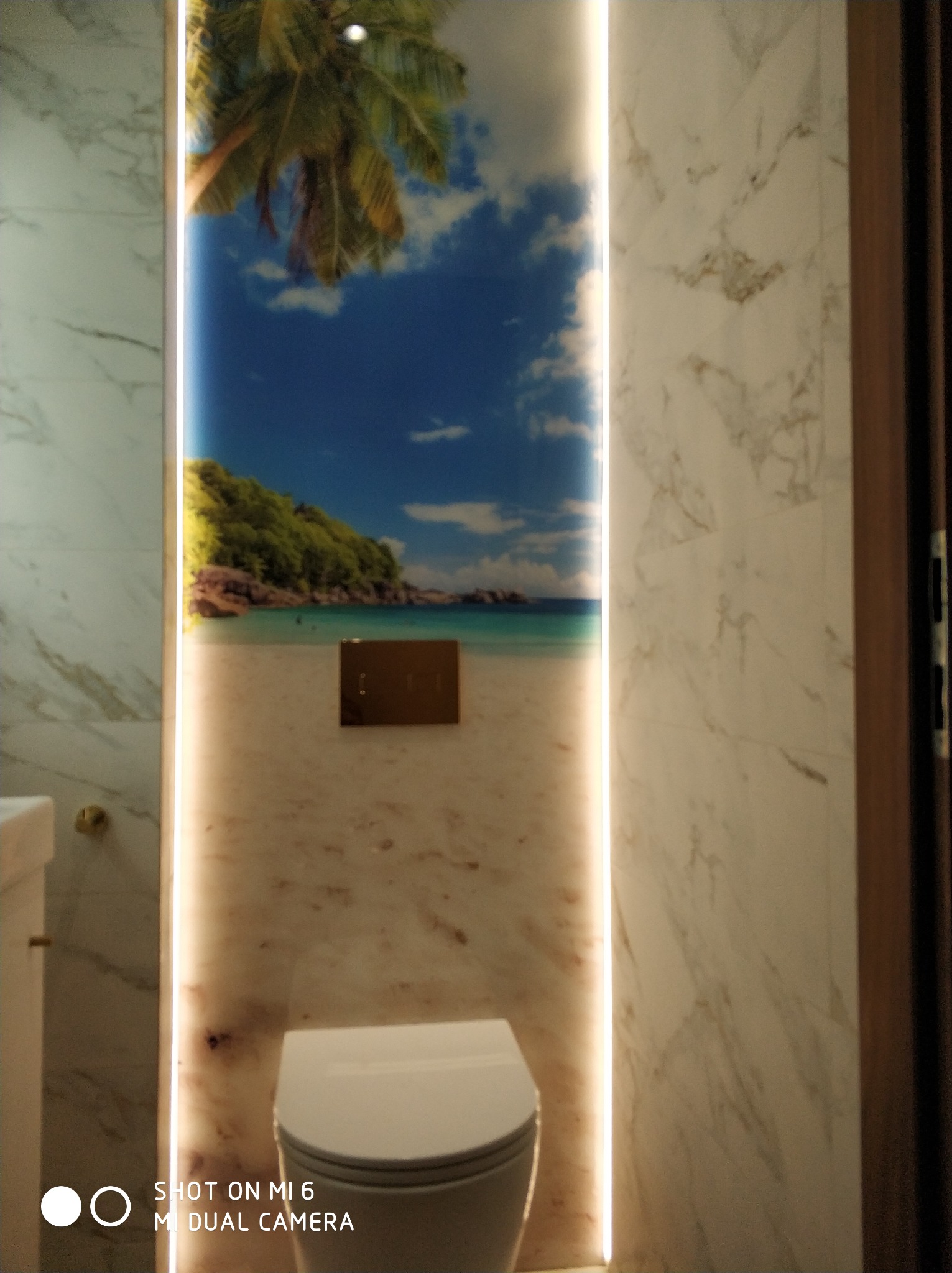 Wykończona łazienka z podświetlaną fototapetą przedstawiającą tropikalną plażę i palmę, nowoczesna toaleta wisząca, marmurowe płytki ścienne.