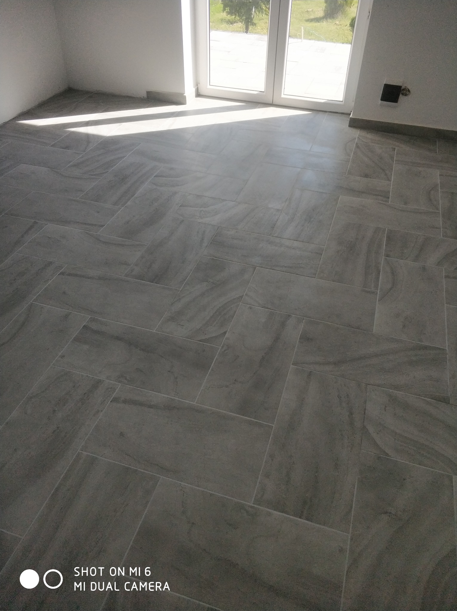 Ułożona pod kątem szara podłoga z płytek imitujących kamień w pustym, jasnym pokoju z dużym oknem balkonowym i wpadającym światłem.