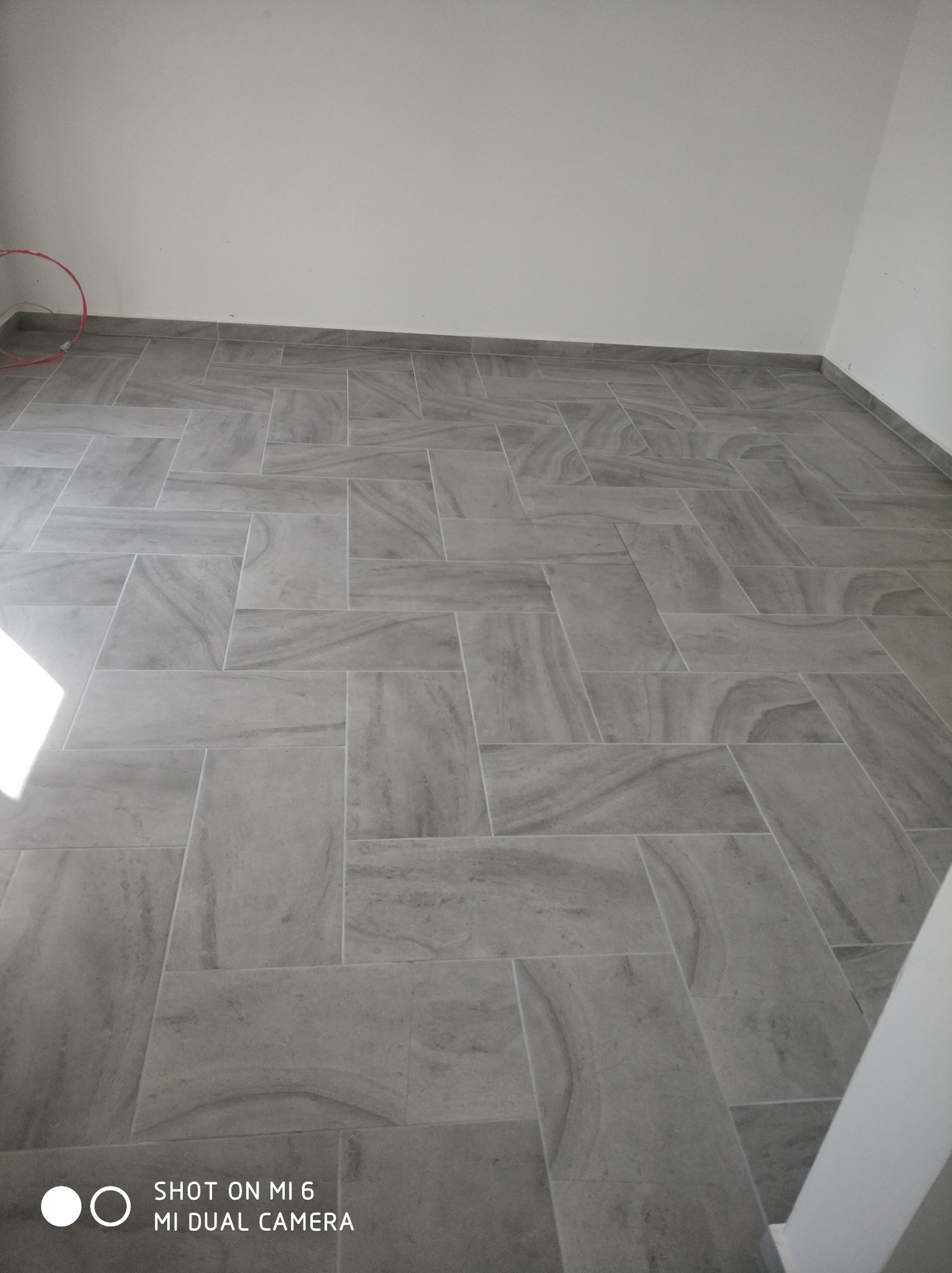 Ułożona szara podłoga z płytek w jodełkę, widoczny fragment białej ściany i rurka instalacyjna przy podłodze.