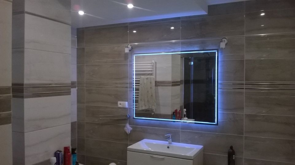 Wykończona łazienka z szarymi płytkami imitującymi kamień, podświetlanym lustrem LED, białą umywalką z szafką i grzejnikiem drabinkowym widocznym w odbiciu.