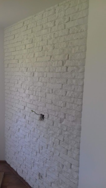 Ściana wyłożona dekoracyjną białą cegłą z widocznym gniazdkiem elektrycznym z wystającymi przewodami, w trakcie prac wykończeniowych.