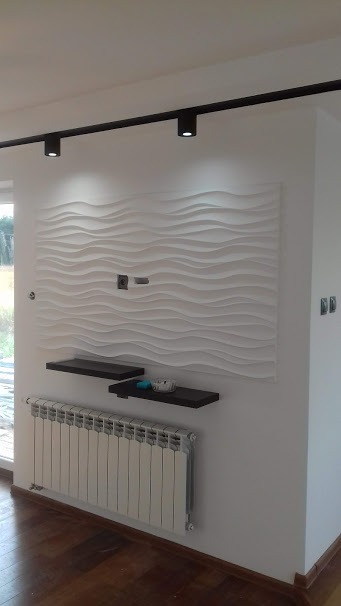 Biała ściana z dekoracyjnym panelem 3D o falistym wzorze, podświetlona czarną listwą z reflektorami, uzupełniona o dwie ciemne półki i grzejnik panelowy.