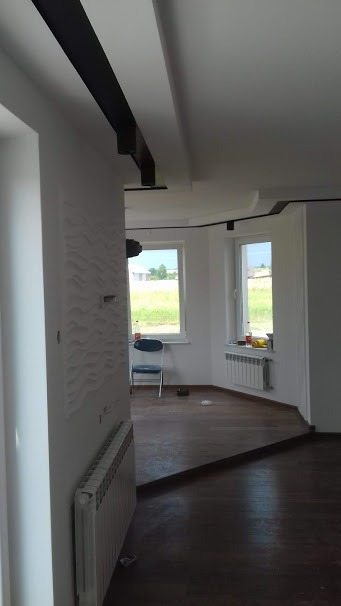 Nowoczesne wnętrze po remoncie: białe ściany z dekoracyjnym panelem 3D, ciemna podłoga z parkietu, czarne listwy sufitowe i oświetlenie punktowe, widok na okna z zielenią na zewnątrz.