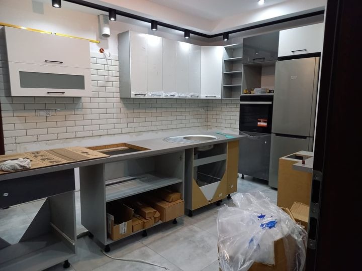 Montaż nowej kuchni: widoczne niedokończone szafki z szarymi blatami, białe płytki na ścianie, srebrna lodówka i piekarnik, oświetlenie sufitowe oraz kartony i folia na podłodze.