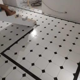 Ułożona podłoga z białych płytek oktagonalnych z czarnymi kwadratami w narożach, obramowana czarną bordiurą, widoczne narzędzia i wiadro po farbie, etap prac wykończeniowych.