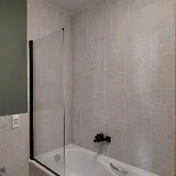 Wykończona łazienka: wanna obudowana płytkami w odcieniach beżu, szklany parawan z czarną ramą, bateria w kolorze czarnym, oświetlenie sufitowe punktowe.