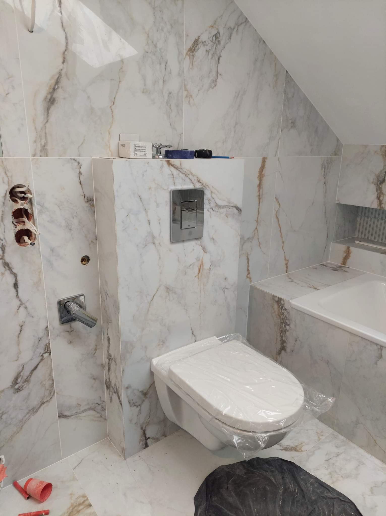 Remont łazienki w trakcie: białe płytki imitujące marmur z brązowymi żyłami, podtynkowa bateria umywalkowa, stelaż WC z przyciskiem, otwory na instalacje elektryczne, zabezpieczona folią toaleta...