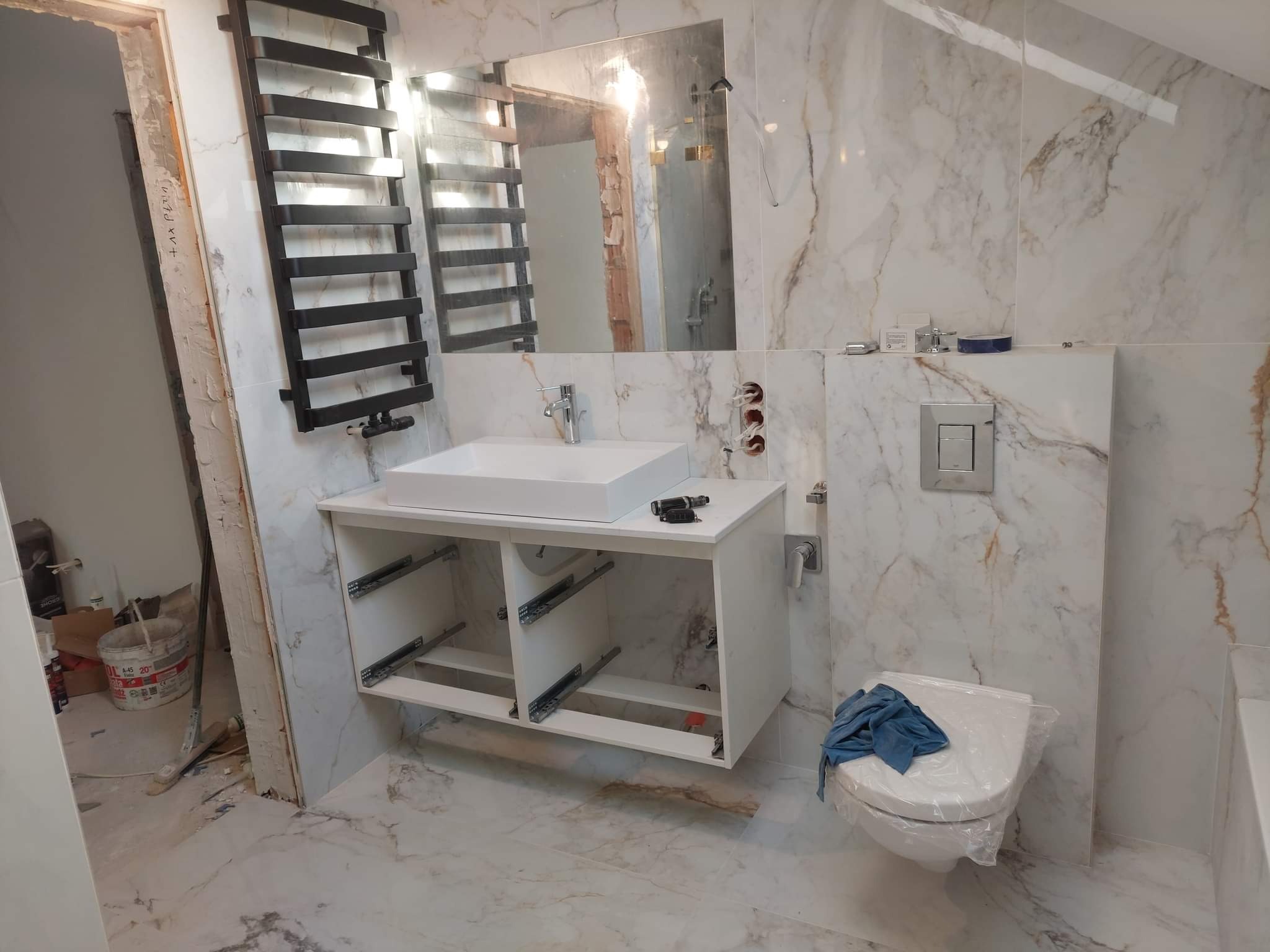 Łazienka w trakcie prac wykończeniowych w Mławie: białe marmurowe płytki, wisząca szafka pod umywalkę z zamontowanymi prowadnicami szuflad, grzejnik drabinkowy i zakryta folią toaleta.