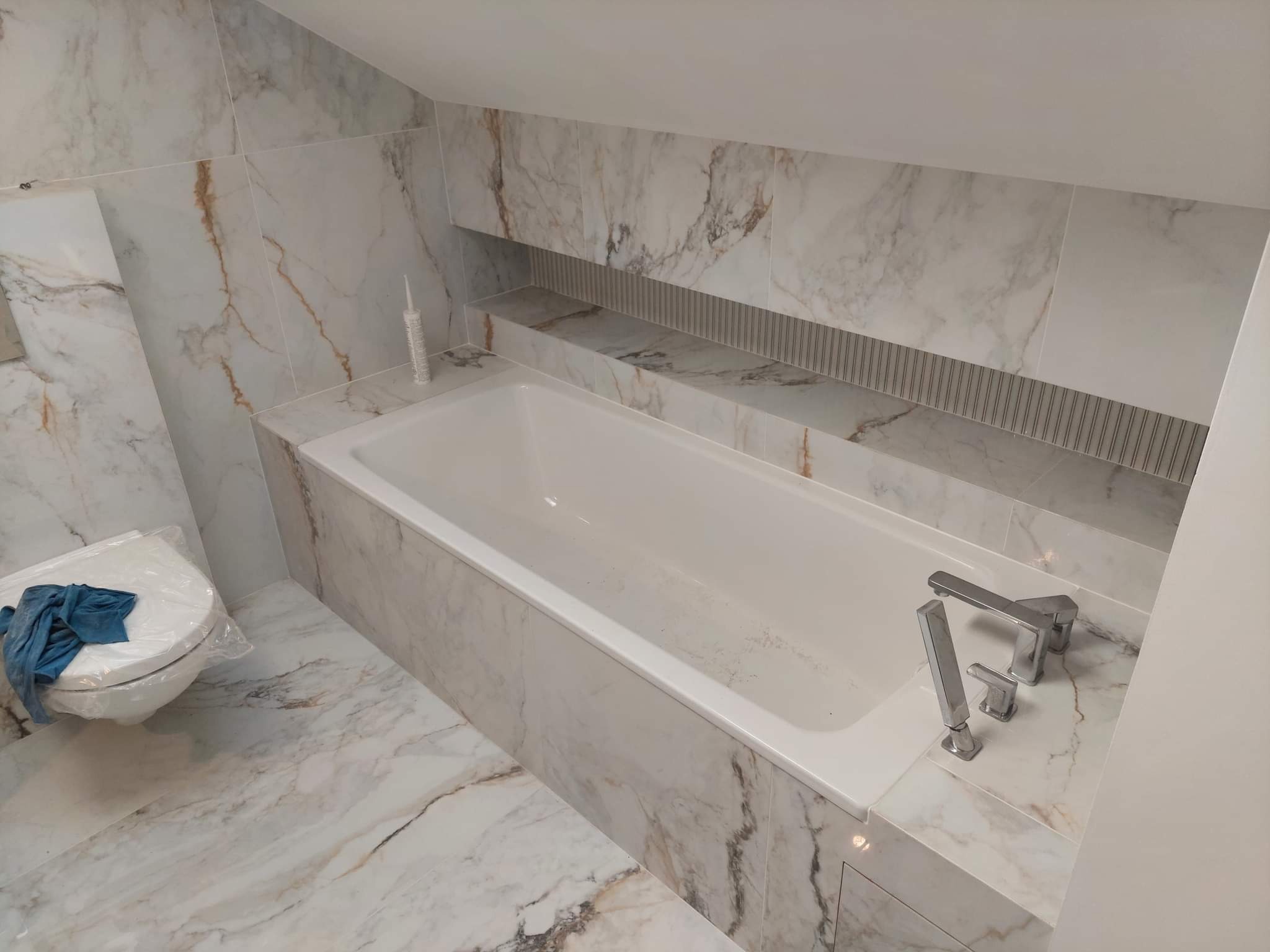 Wykończona łazienka z wanną obudowaną marmurowymi płytkami, armaturą i zabezpieczonym folią sedesem, widoczny tubka silikonu.