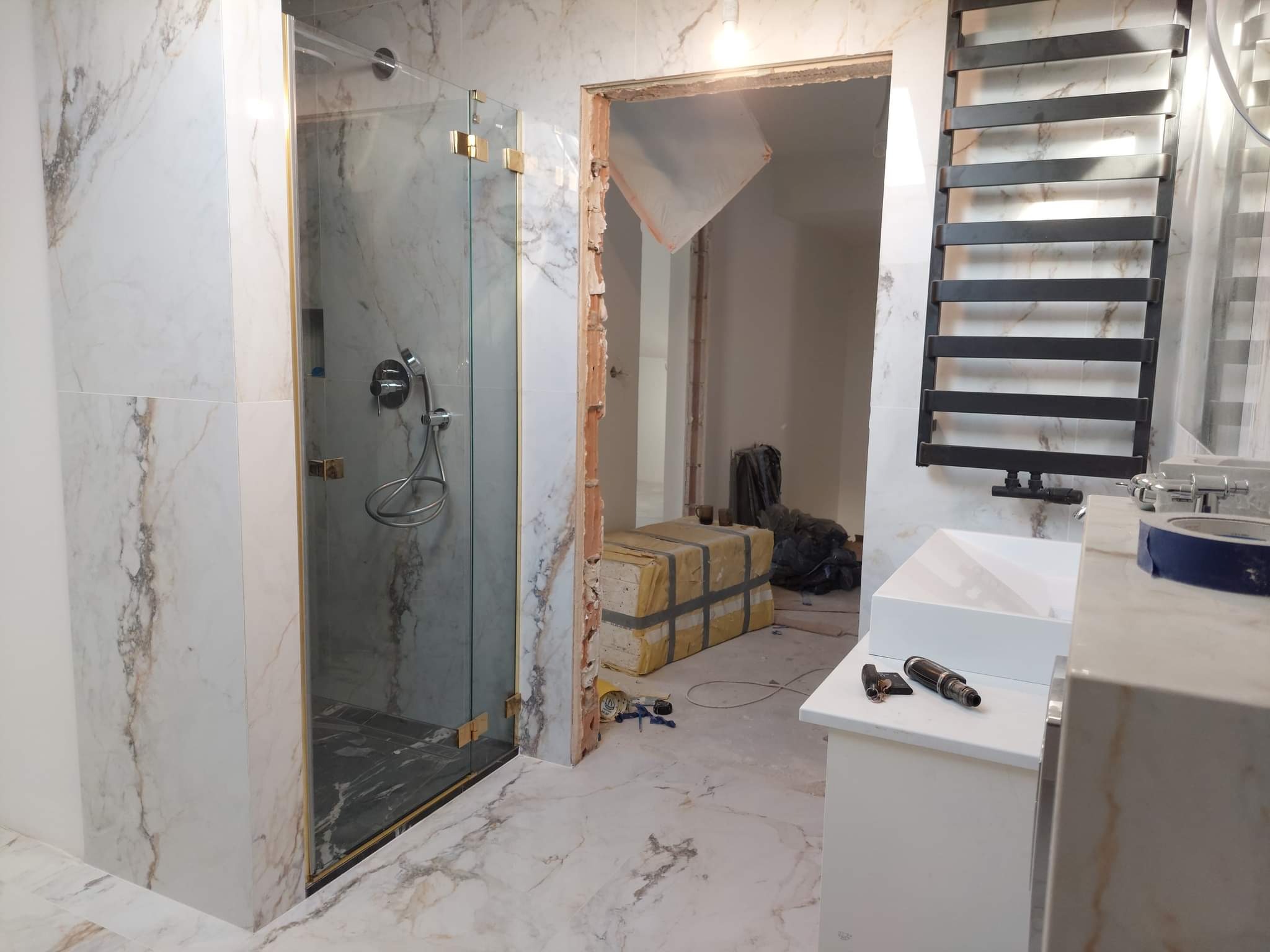 Łazienka w trakcie prac: kabina prysznicowa ze złotymi zawiasami, marmurowe płytki na ścianach i podłodze, grzejnik drabinkowy, umywalka z baterią, narzędzia na blacie, materiały budowlane...