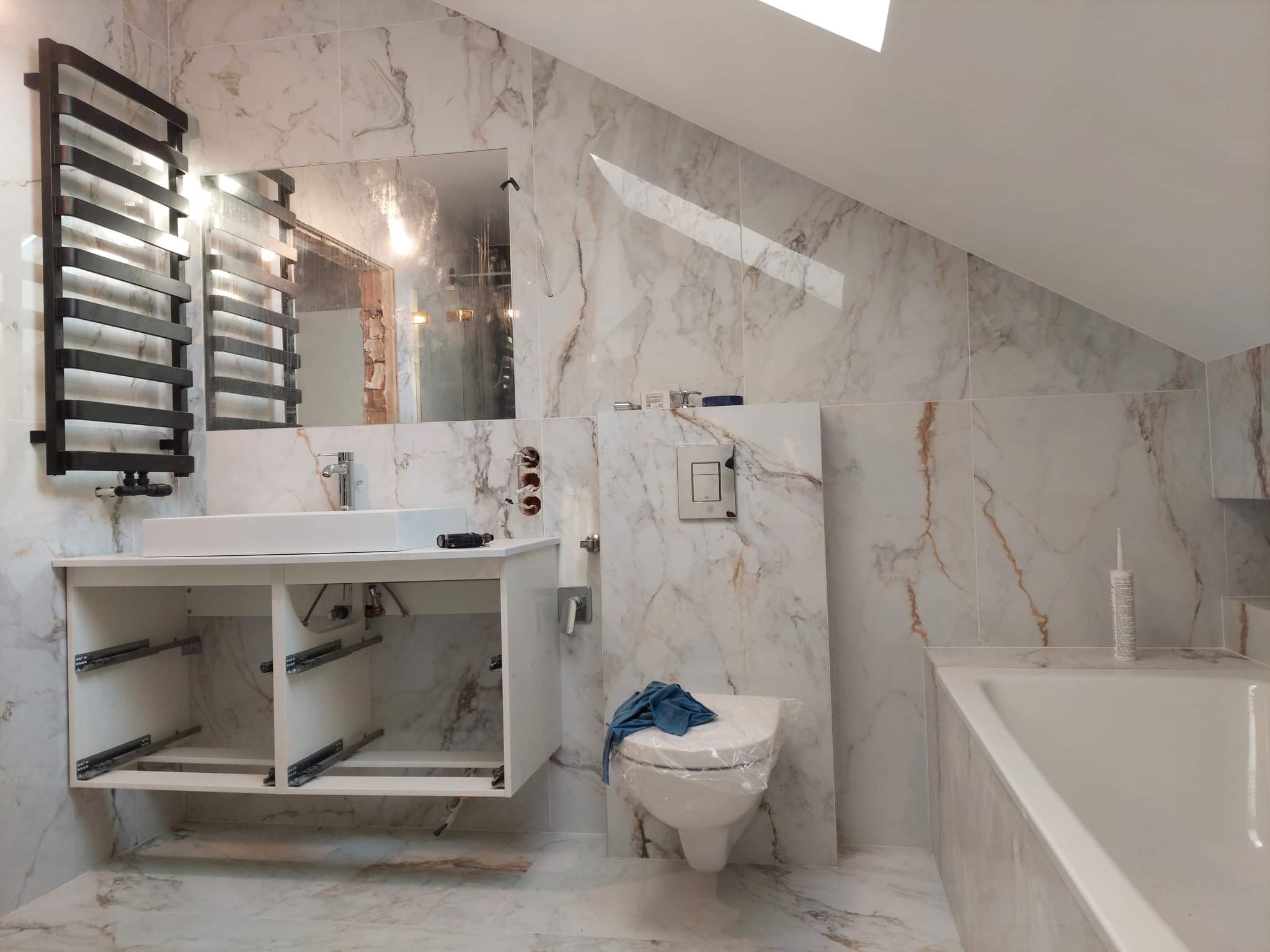 Łazienka w trakcie montażu: białe marmurowe płytki, wisząca szafka pod umywalkę z otwartymi półkami, toaleta z niebieską szmatką, grzejnik drabinkowy i wanna, częściowo wykończone ściany.