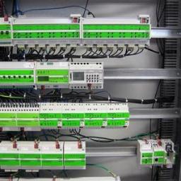 Nowoczesne Instalacje Elektryczne - Rozdzielnica elektryczna z modułowymi zabezpieczeniami i sterownikami, widoczne okablowanie i szyny montażowe, elementy w kolorze białym i zielonym.