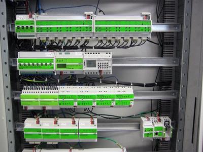 Rozdzielnica elektryczna z modułowymi zabezpieczeniami i sterownikami, widoczne okablowanie i szyny montażowe, elementy w kolorze białym i zielonym.