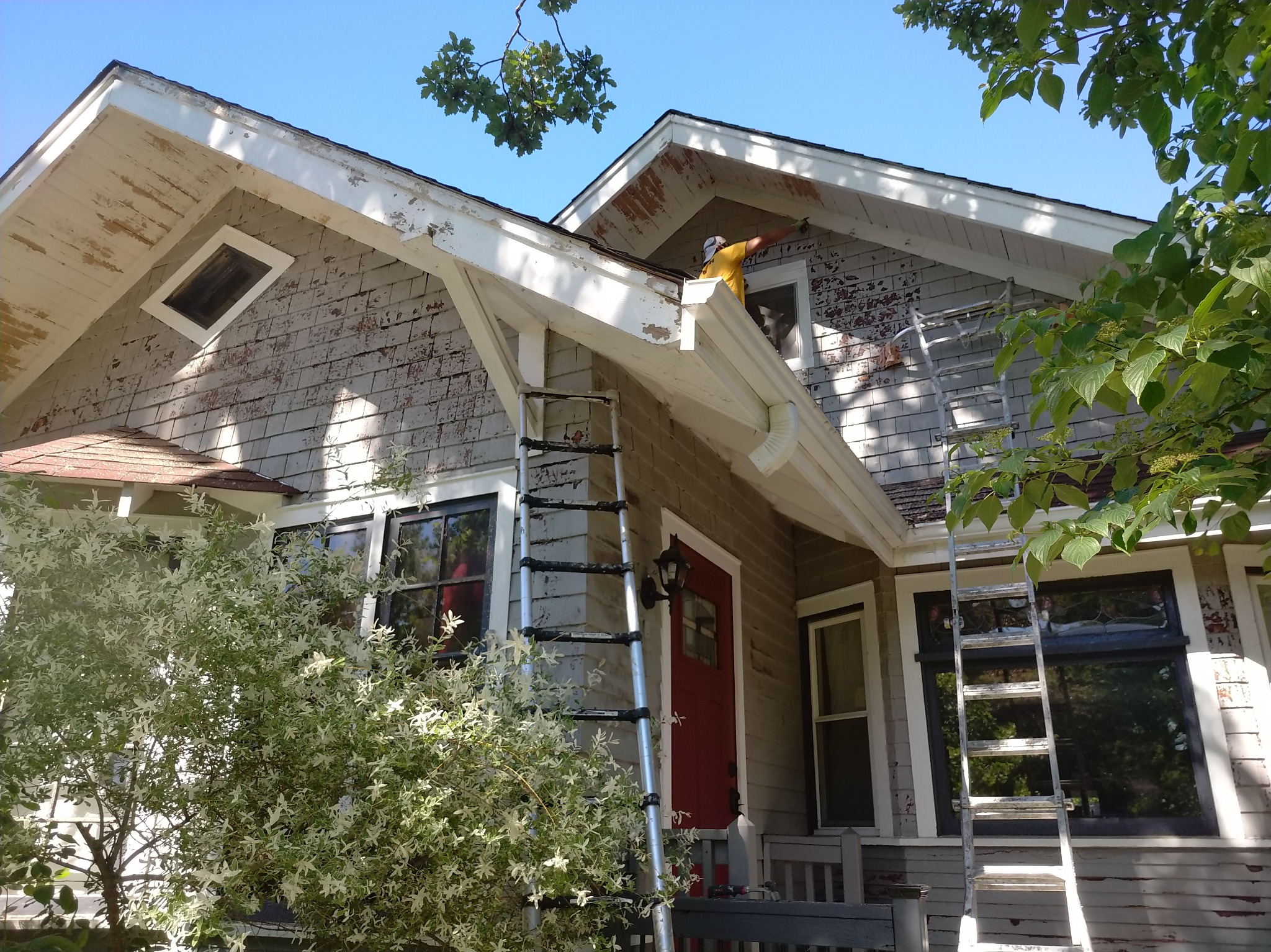 Pracownik w żółtej koszulce maluje elewację domu z drabiny, widoczne łuszczące się fragmenty starej farby i odrapane drewniane elementy.