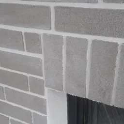 Szara ściana z cegły klinkierowej z białymi fugami wokół okna z czarną ramą, zabezpieczonego folią ochronną.