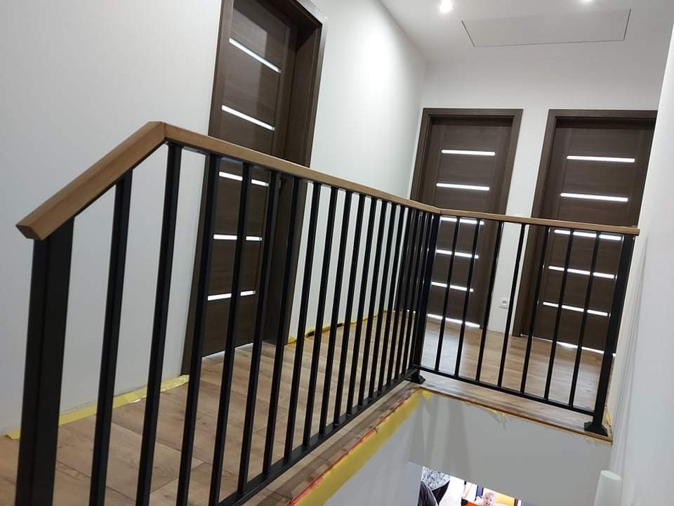 Nowoczesna balustrada schodowa z czarnych metalowych prętów i drewnianym poręczą, trzy drzwi z poziomymi przeszkleniami w tle.