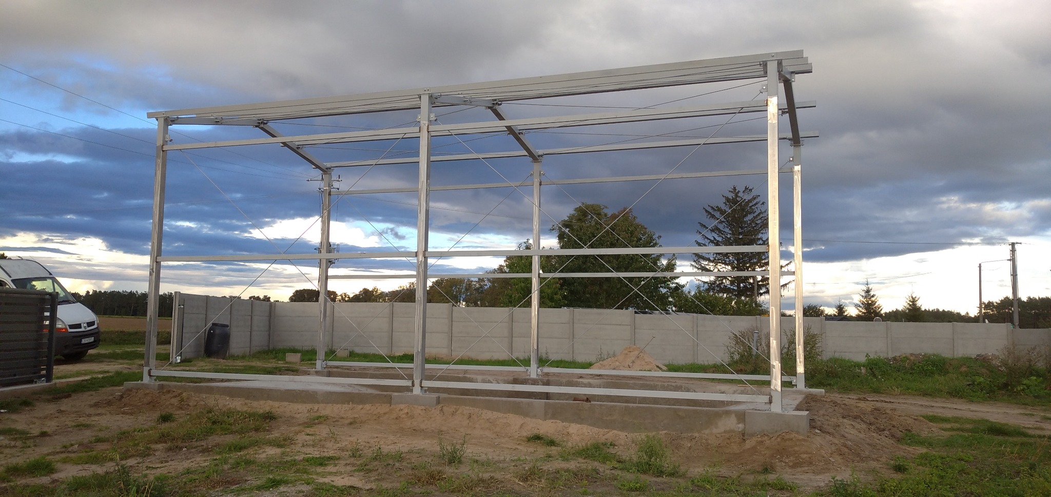 Metalowa konstrukcja z profili, prawdopodobnie zadaszenie, w trakcie budowy, na betonowej podstawie, w tle betonowy płot i zachmurzone niebo.