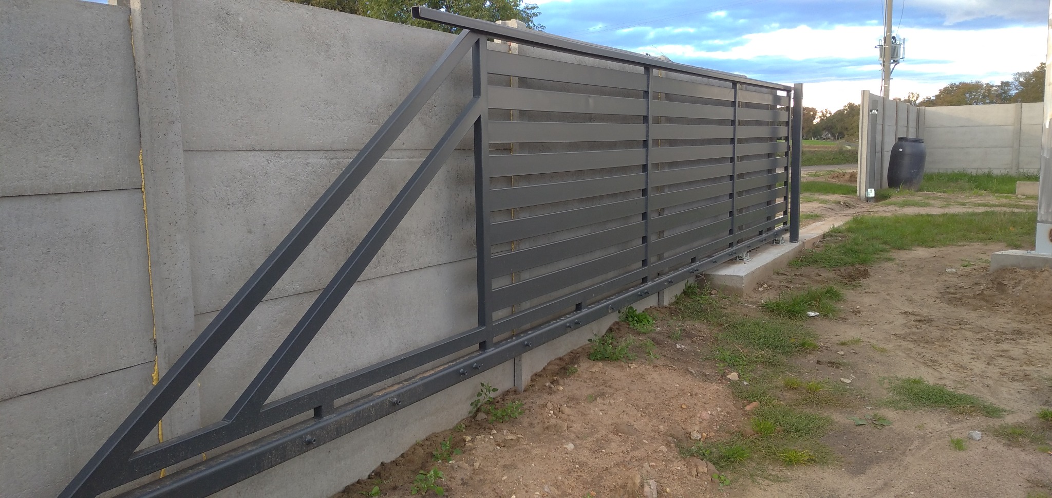 Nowoczesna, przesuwna brama wjazdowa z poziomych, szarych profili metalowych, zamontowana na betonowym fundamencie i przylegająca do betonowego muru z widocznymi łączeniami płyt.