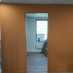 Lukasz Witek - Pomarańczowe ściany w biurze po malowaniu, widok przez otwarte drzwi na jasne pomieszczenie z oknem, grzejnikiem i narzędziami, podłoga wyłożona panelami.