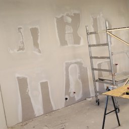 Lukasz Witek - Ściana w trakcie szpachlowania z widocznymi łatami gipsowymi, drabiny malarskie, stół roboczy z narzędziami: miarka, kombinerki, oświetlenie budowlane.