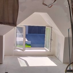 Lukasz Witek - Poddasze w trakcie prac wykończeniowych, widoczne okno dachowe z otwartymi skrzydłami, świeżo pomalowane ściany na biało, zabezpieczone elementy konstrukcyjne folią, oraz czarny plecak leżący...