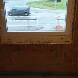 Lukasz Witek - Widok z okna na ulicę z przejeżdżającym samochodem, okno osadzone w surowej, ceglanej ścianie, widoczne ślady pianki montażowej.