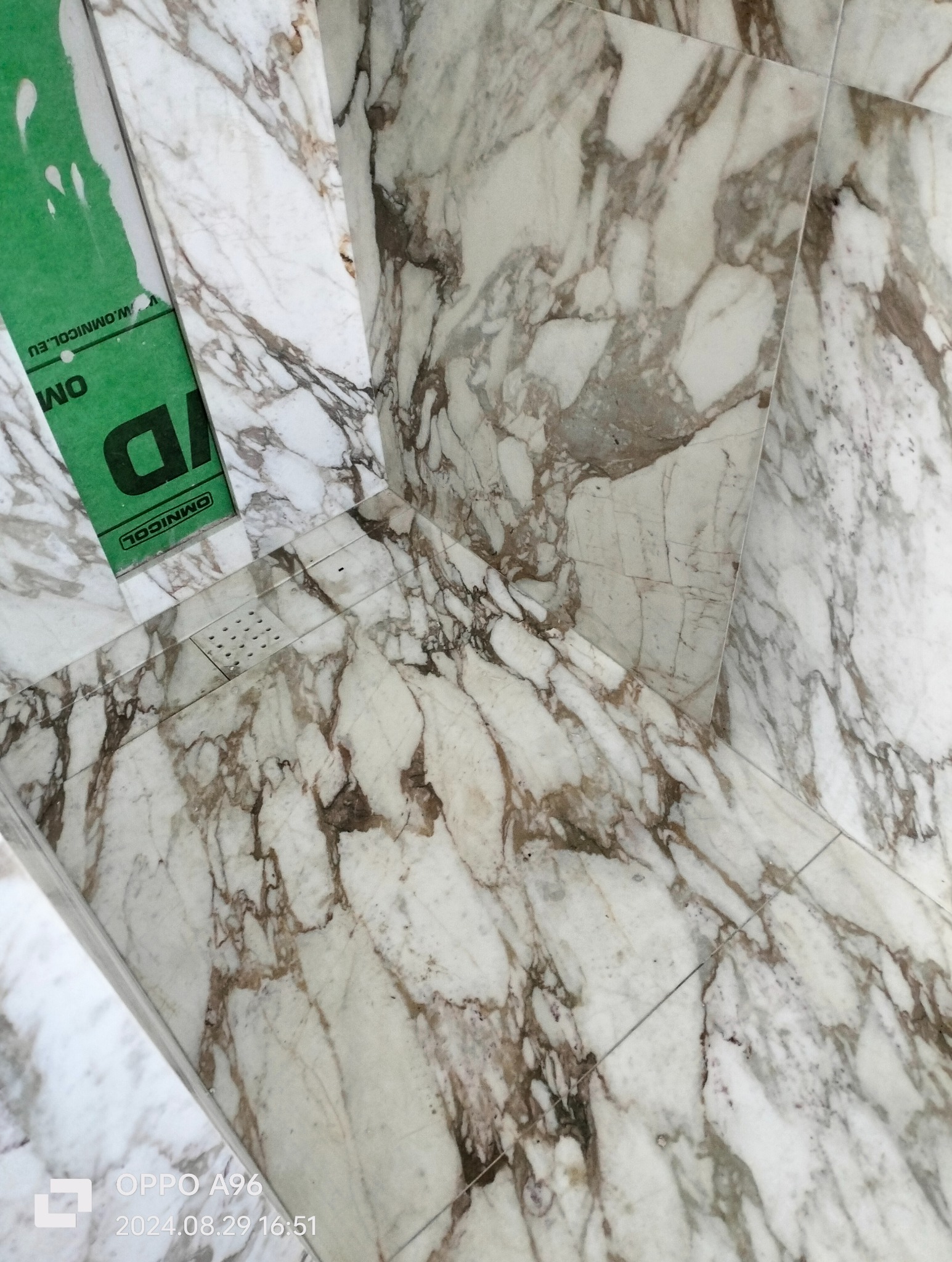 Fragment wykończonego brodzika z płytek imitujących marmur, widoczny odpływ liniowy oraz fragment zielonej taśmy zabezpieczającej.