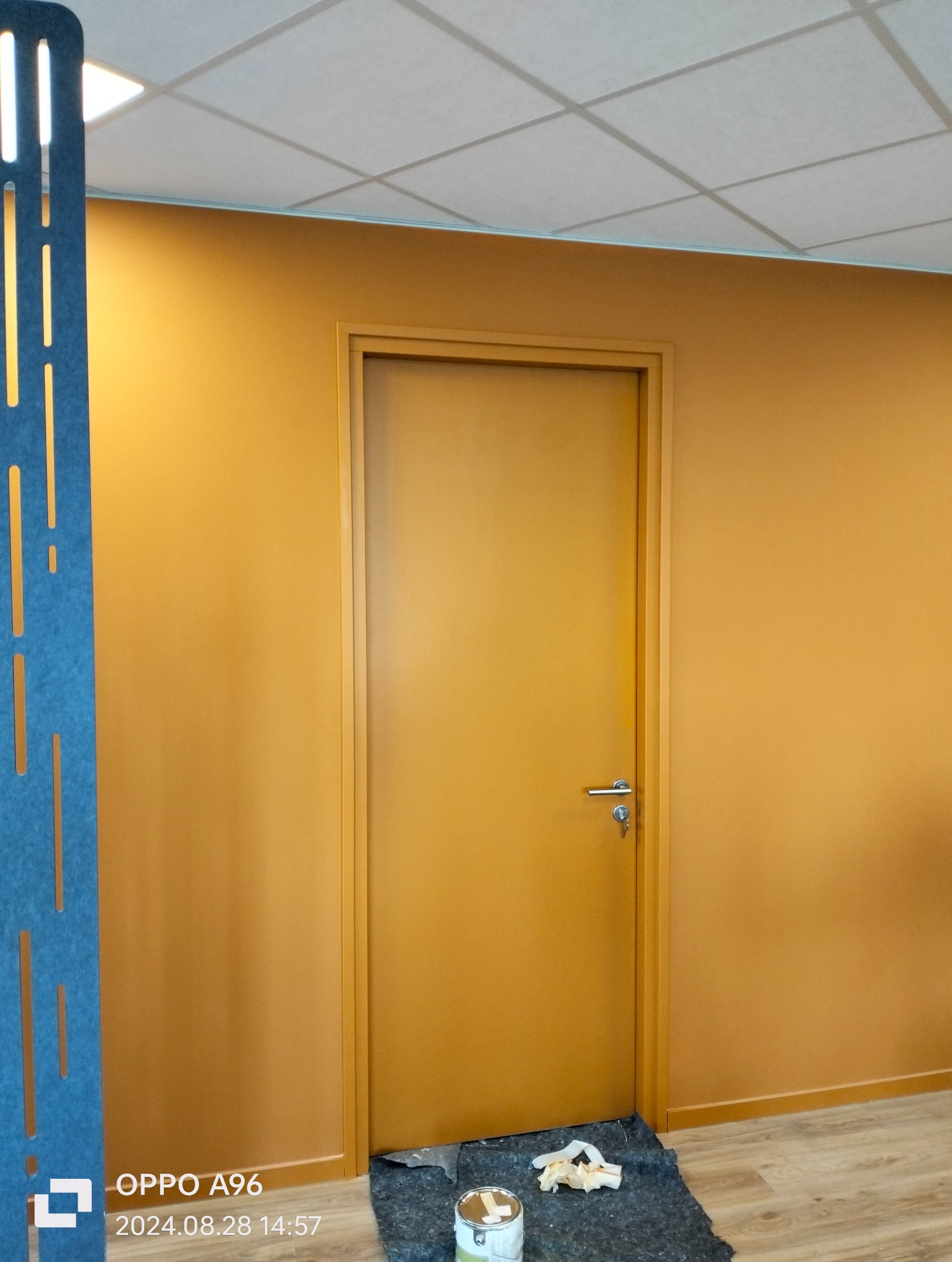 Świeżo pomalowane biuro z żółtymi ścianami i dopasowaną kolorystycznie stolarką drzwiową; widoczna puszka farby i zabezpieczenie podłogi.