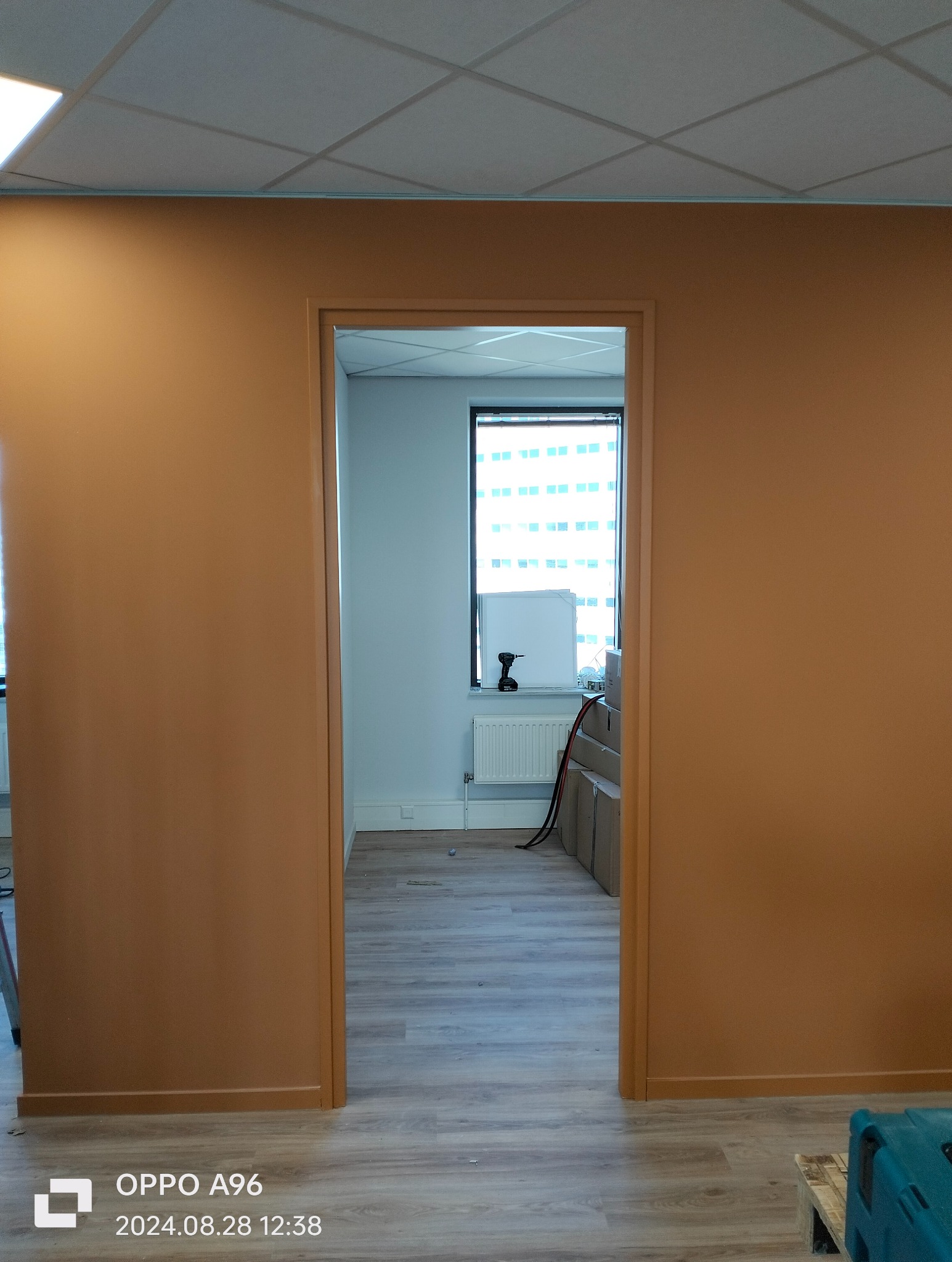 Pomarańczowe ściany w biurze po malowaniu, widok przez otwarte drzwi na jasne pomieszczenie z oknem, grzejnikiem i narzędziami, podłoga wyłożona panelami.