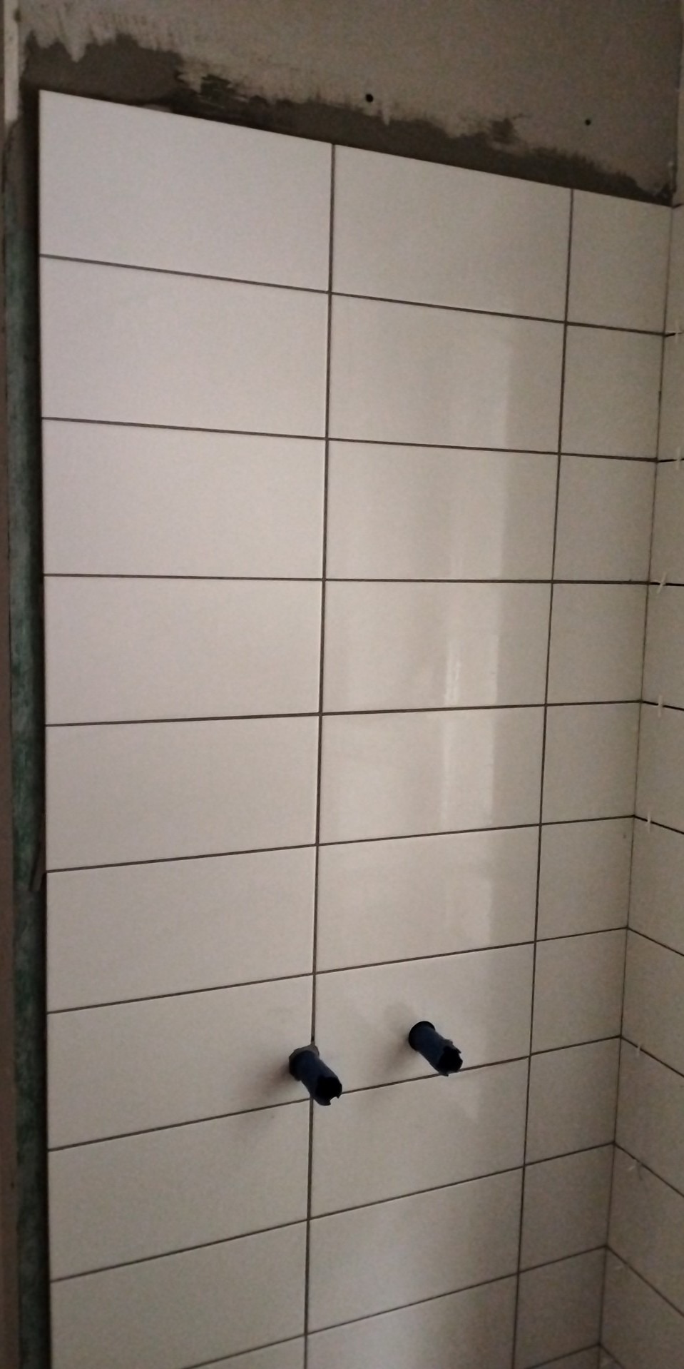 Ściana z ułożonymi białymi, prostokątnymi płytkami, widoczne czarne fugi i dwa otwory z niebieskimi osłonkami na rury, ślady zaprawy na górze ściany.