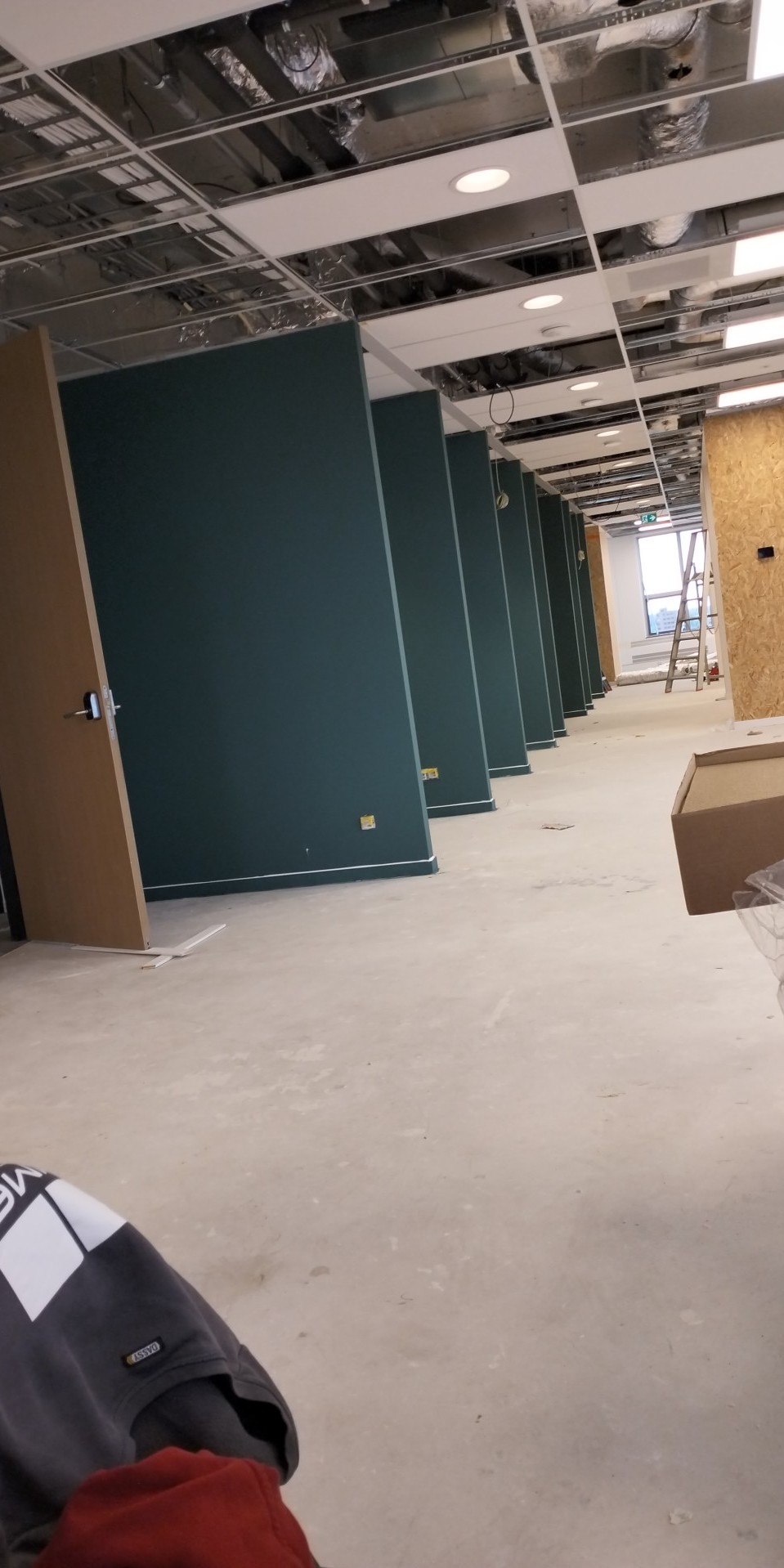 Wnętrze biura w trakcie remontu: świeżo pomalowane na ciemnozielono ściany działowe, sufit podwieszany z widoczną instalacją, częściowo zamontowane drzwi, drabina w tle przy oknie.