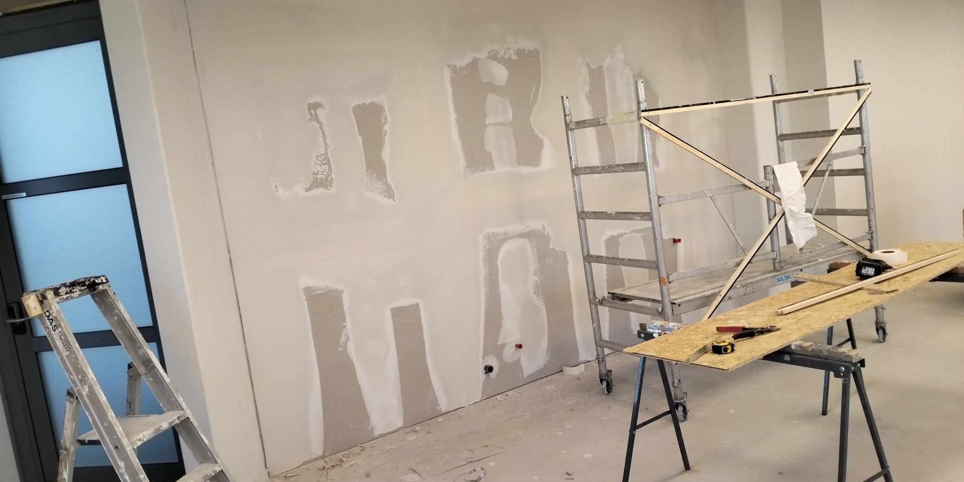 Ściana w trakcie szpachlowania z widocznymi łatami gipsowymi, drabiny malarskie, stół roboczy z narzędziami: miarka, kombinerki, oświetlenie budowlane.