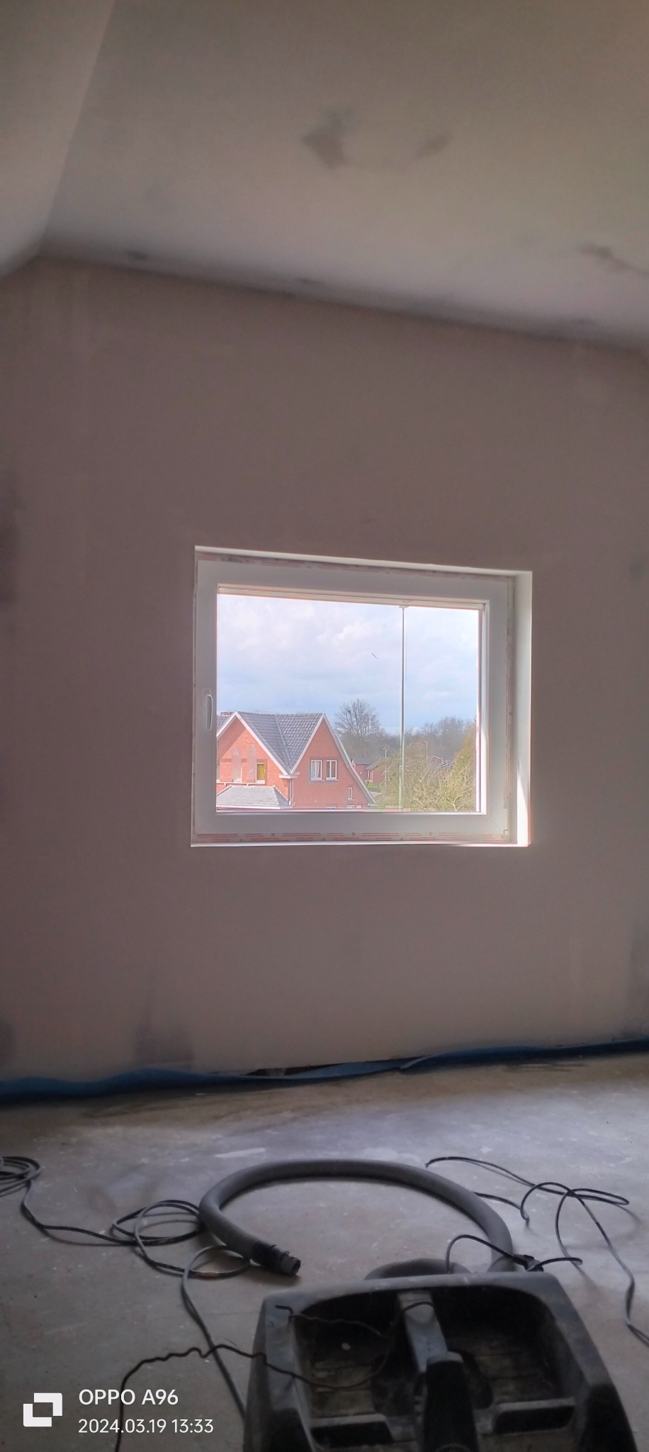 Pokój w trakcie prac wykończeniowych, ściany po nałożeniu gładzi, widoczny odkurzacz przemysłowy i zabezpieczenie podłogi, w tle widok na dom przez okno.