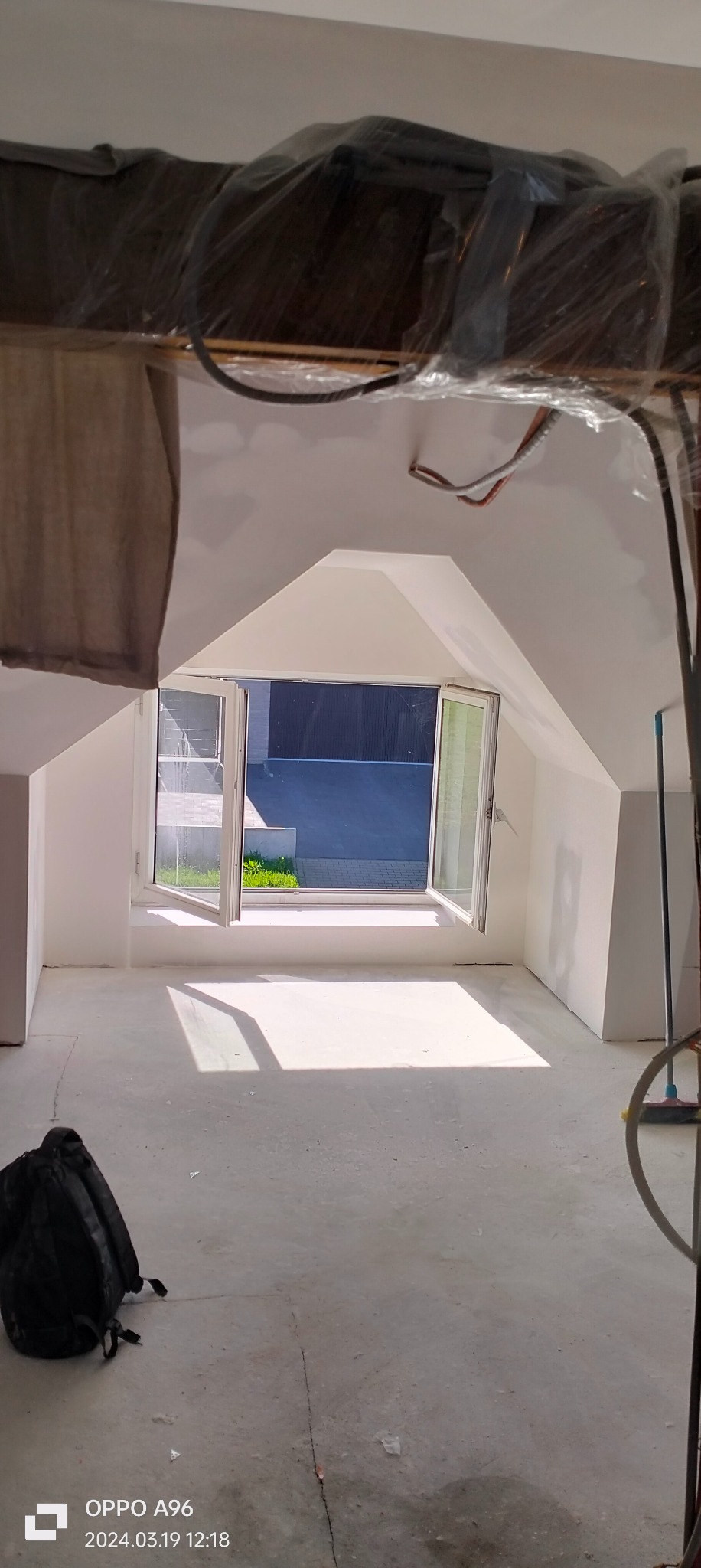 Poddasze w trakcie prac wykończeniowych, widoczne okno dachowe z otwartymi skrzydłami, świeżo pomalowane ściany na biało, zabezpieczone elementy konstrukcyjne folią, oraz czarny plecak leżący...