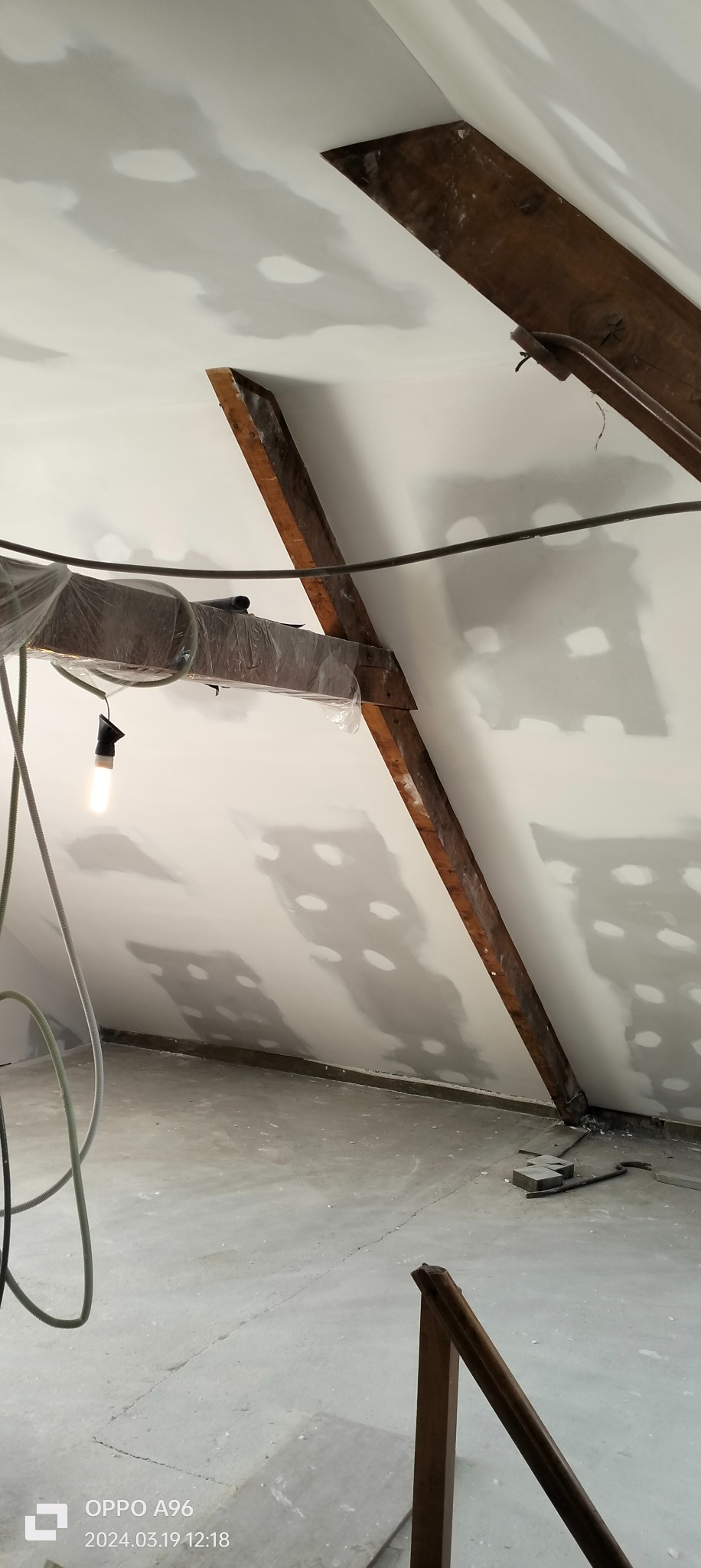 Poddasze w trakcie prac wykończeniowych: drewniane belki konstrukcyjne, częściowo osłonięte folią, biała ściana z widocznymi punktowymi miejscami po szpachlowaniu, surowa żarówka na kablu, betonowa...