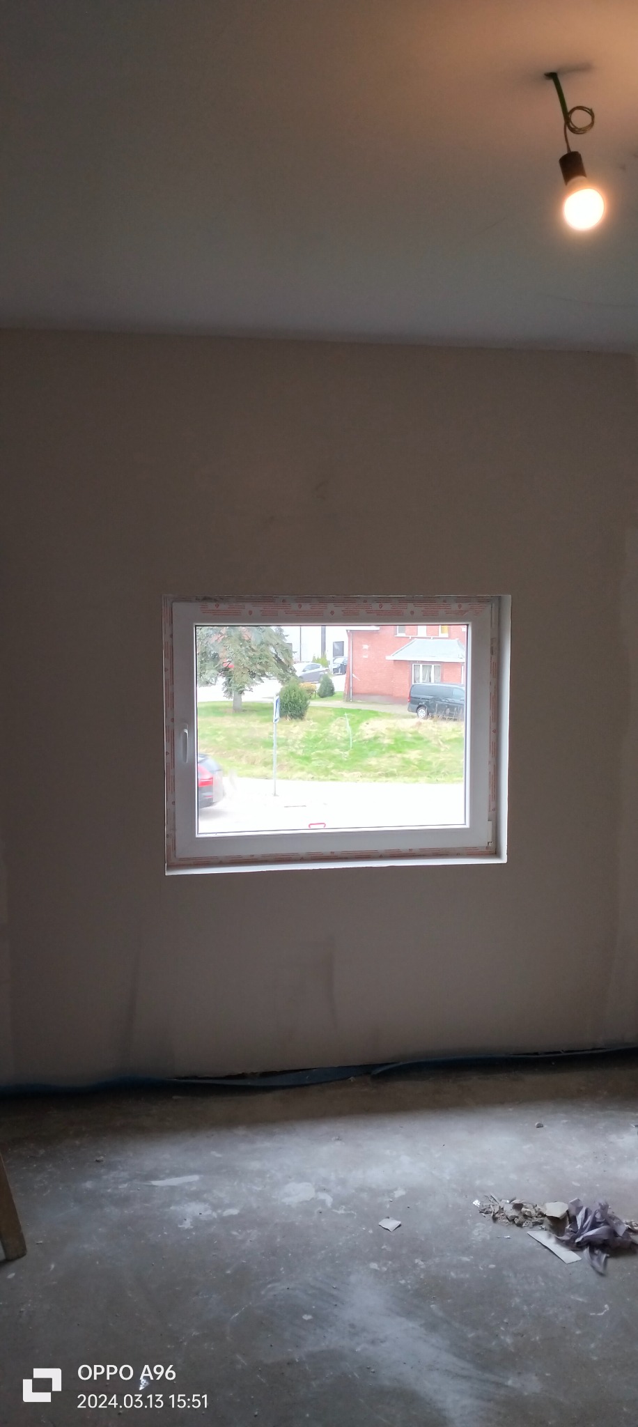 Pokój w trakcie prac wykończeniowych: ściany po nałożeniu gładzi, widoczne nierówności i ślady po szpachlowaniu, okno z widokiem na zewnątrz, zabezpieczona podłoga, surowa żarówka na suficie.