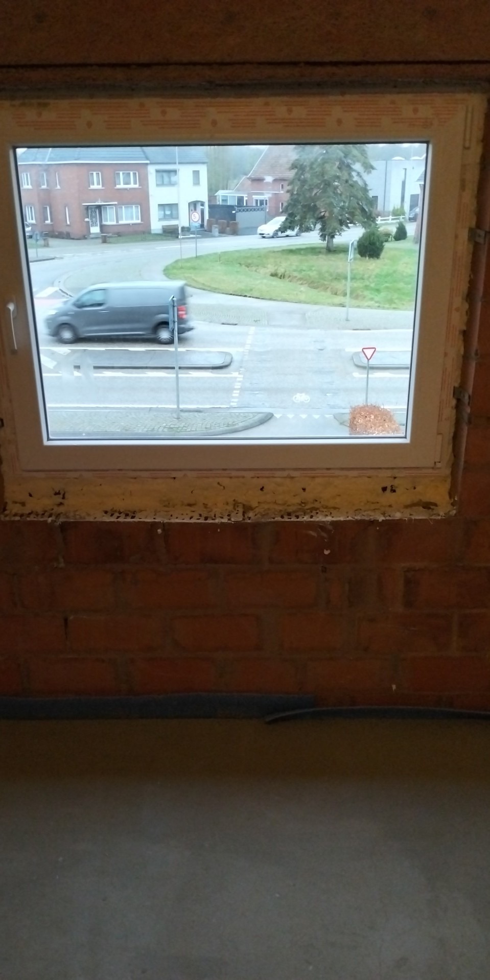 Widok z okna na ulicę z przejeżdżającym samochodem, okno osadzone w surowej, ceglanej ścianie, widoczne ślady pianki montażowej.