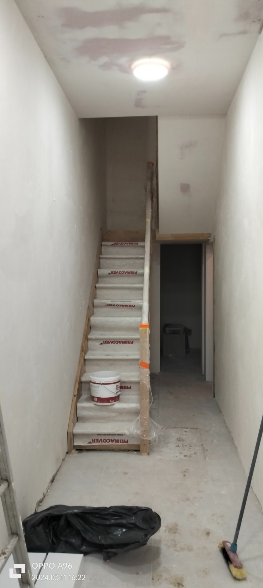 Klatka schodowa w trakcie przygotowań do malowania: schody zabezpieczone folią i kartonem PRIMACOVER, ściany częściowo wyszpachlowane, widoczne ślady po szpachlowaniu na suficie, wiadro farby...