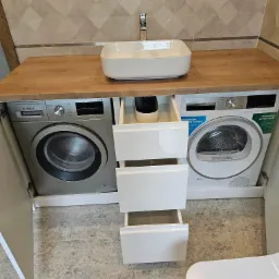 Łazienka z drewnianym blatem, na którym umieszczono nablatową, białą umywalkę. Pod blatem zabudowane pralka i suszarka firmy Bosch, pomiędzy nimi białe szuflady z otwartymi frontami. Nad blatem...