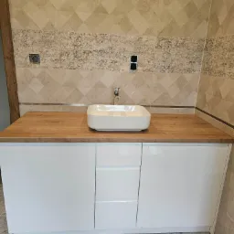 Nowoczesna łazienka z białą szafką pod umywalkę z trzema szufladami, drewniany blat, prostokątna umywalka nablatowa, neutralne płytki na ścianach i podłodze, widoczne gniazdka elektryczne.