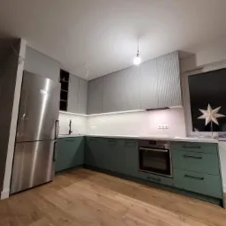 Nowoczesna kuchnia w odcieniach szarości i zieleni z drewnianą podłogą, oświetleniem LED pod szafkami górnymi i wiszącą żarówką bez klosza.