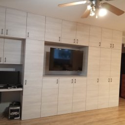 Lili Meble - Ireneusz Figiel - Jasna zabudowa meblowa z szafkami i wnęką na telewizor. Po lewej stronie biurko z komputerem i sprzętem audio. Widoczny parkiet i sufit z wentylatorem.