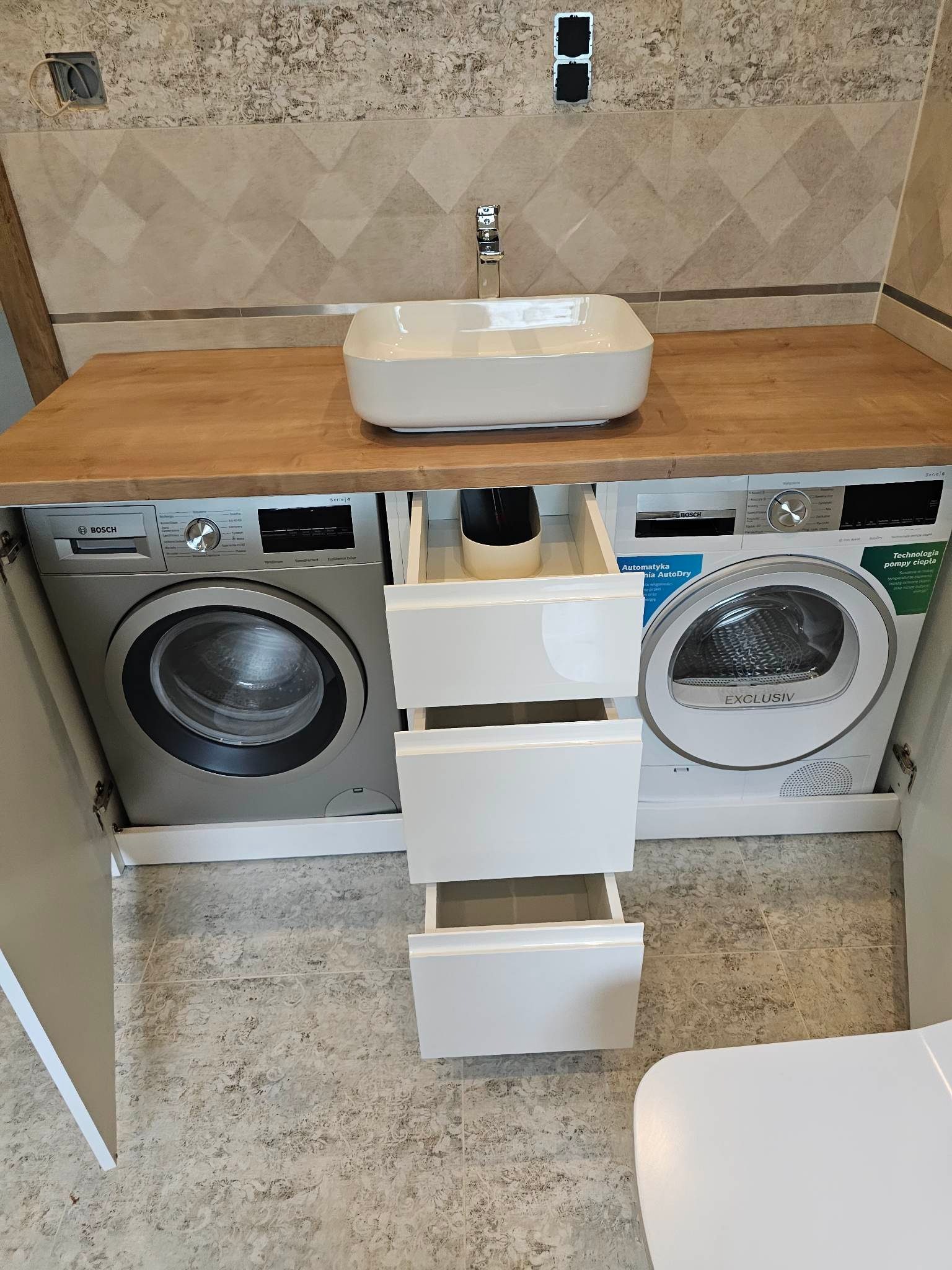 Łazienka z drewnianym blatem, na którym umieszczono nablatową, białą umywalkę. Pod blatem zabudowane pralka i suszarka firmy Bosch, pomiędzy nimi białe szuflady z otwartymi frontami. Nad blatem...